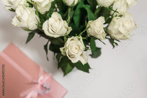 White tea roses stand in a glass vase  which is tied with a red ribbon and stands on the table. There is a gift nearby. in a pink box. Gift bouquet