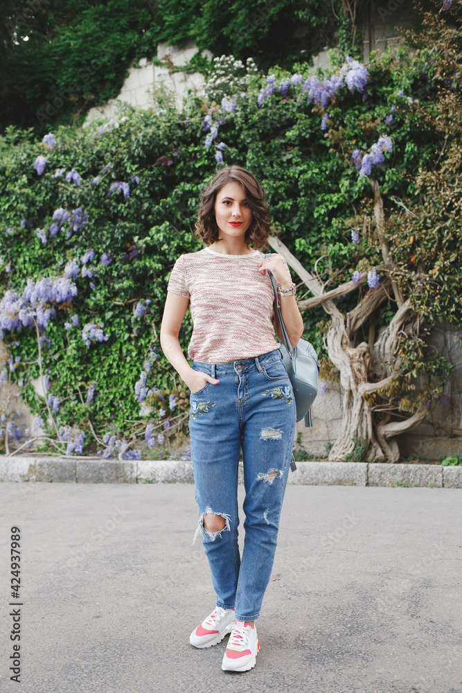 Young beautiful woman wearing jeans, pink tee shirt, sneakers and backpack walking near a stone wall covered with flowering climbing purple Wisteria and green ivy leaves. Spring or summer portrait Young beautiful woman wearing jeans, pink tee shirt, sneakers and backpack walking near a stone wall covered with flowering climbing purple Wisteria and green ivy leaves. Spring or summer portrait
