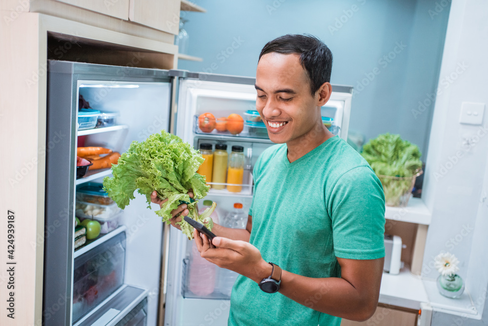 happy smiling man ordering online groceries shop. man standing in front of the fridge while holding his mobile phone happy smiling man ordering online groceries shop. man standing in front of the fridge while holding his mobile phone