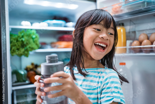 thirsty happy young asian girl open fridge door drinking a bottle of water photo