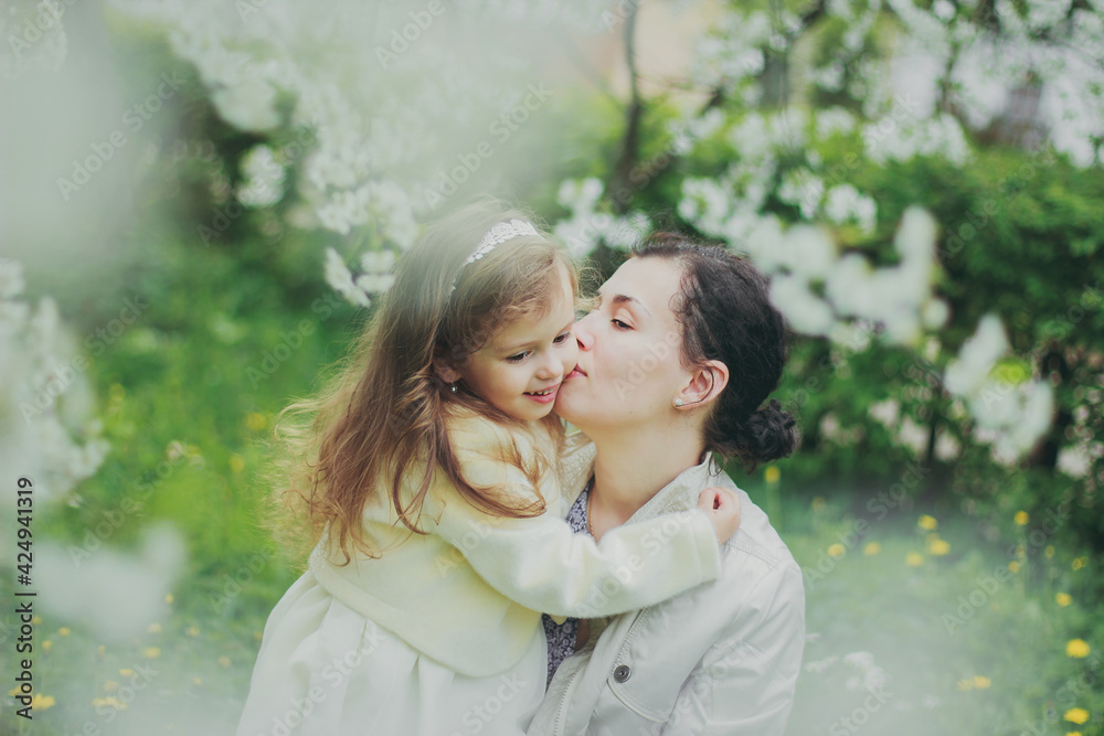 Fototapeta premium Happy mother's day. Little girl hugs her mother in the spring cherry garden. Portrait of happy mother and daughter among white flowers trees. Family values. Motherhood. Mom and child in blooming park.