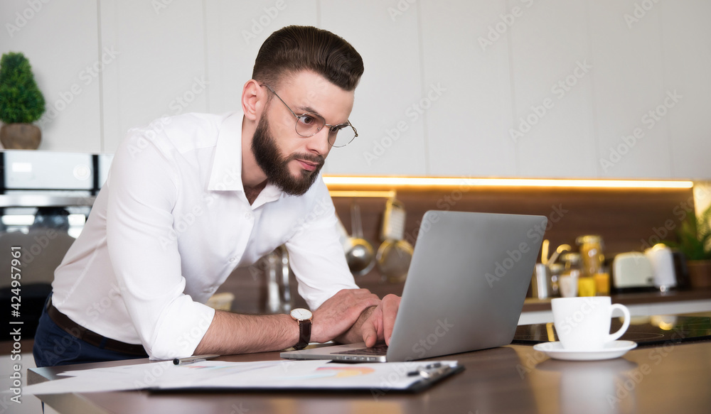 Handsome confident businessman in smart casual wear is working with laptop at domestic kitchen Handsome confident businessman in smart casual wear is working with laptop at domestic kitchen