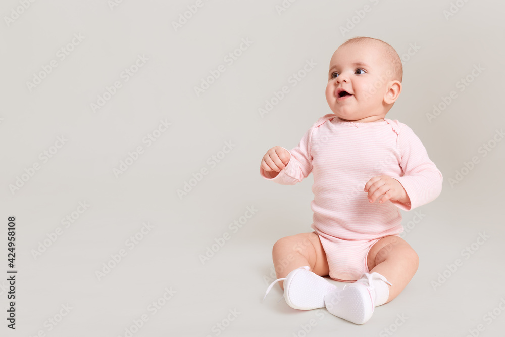 Happy excited infant child baby girl toddler wearing bodysuit and socks sitting on floor isolated on white background looking away with open mouth at free copy space. Happy excited infant child baby girl toddler wearing bodysuit and socks sitting on floor isolated on white background looking away with open mouth at free copy space.