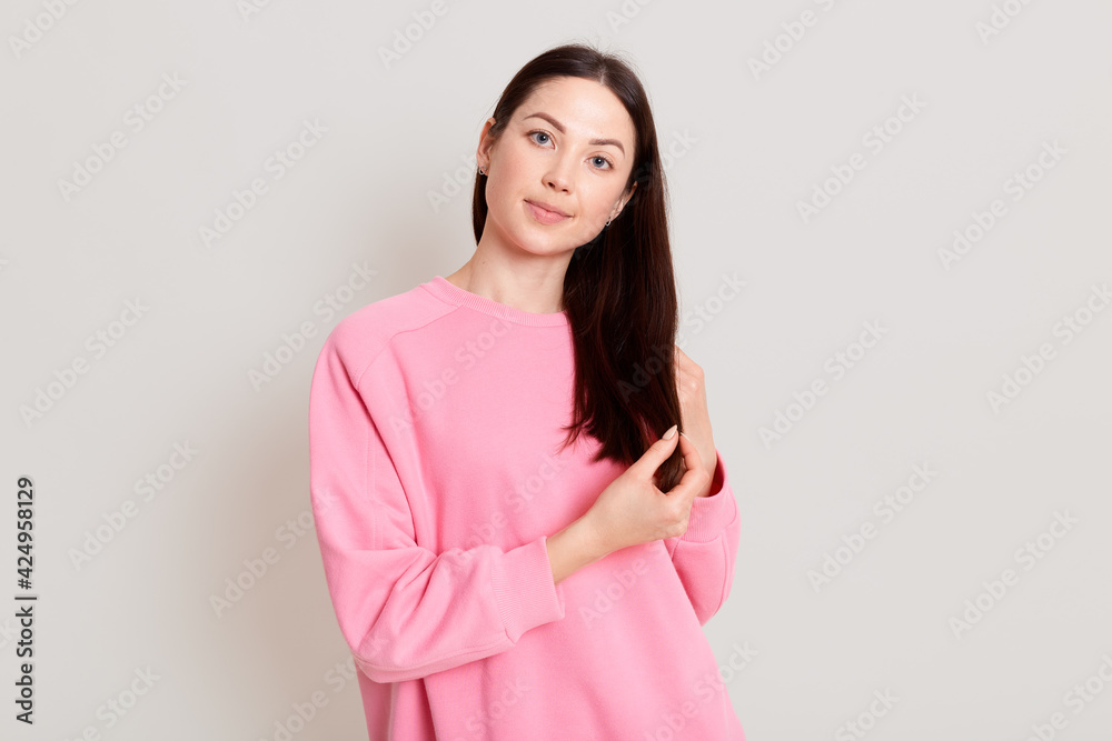 Portrait of charming dark haired female wearing pink sweatshirt, looking at camera with calm expression and cute smile, touching her hair with hands, isolated over white background. Portrait of charming dark haired female wearing pink sweatshirt, looking at camera with calm expression and cute smile, touching her hair with hands, isolated over white background.