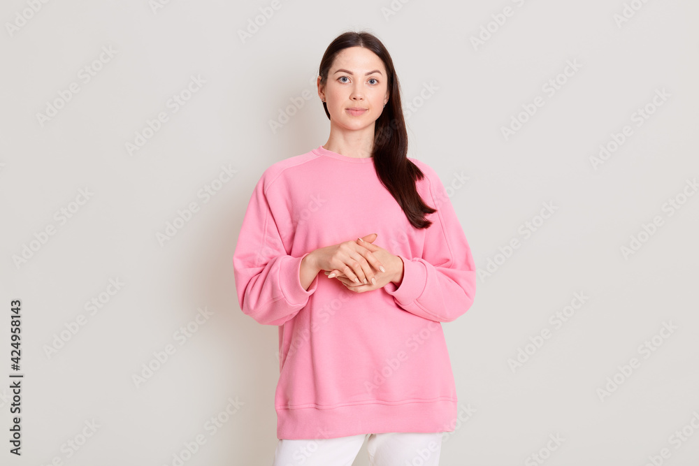 Pretty calm female with dark hair, dressed casual pants and pink sweatshirt, looking with calm expression at camera, keeps hands together in front of chest. Caucasian girl isolated on light background Pretty calm female with dark hair, dressed casual pants and pink sweatshirt, looking with calm expression at camera, keeps hands together in front of chest. Caucasian girl isolated on light background