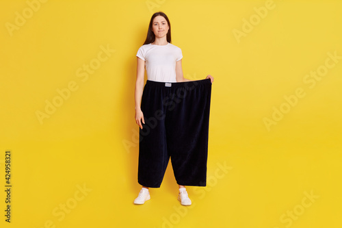 Full length photo of Young adult Caucasian girl wearing casual t shirt and too big size pants, looks at camera with puzzled expression and bewilderment, purses lips, isolated over yellow background. photo