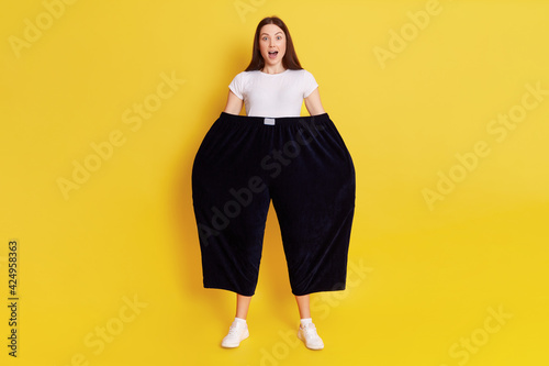 Shocked astonished woman wearing old too big black trousers keeps hands in pants. looks at camera with open mouth and big eyes, has surprised facial expression, posing isolated over yellow background. photo