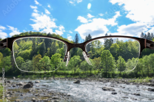 River and green forest landscape focused in women's glasses frame. View through eyeglasses. Better vision concept. Colorful view of sunset landscape in glasses.