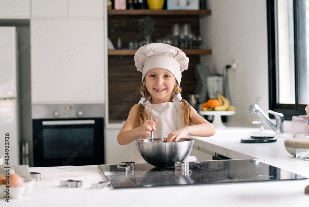 Small cute girl in special clothes and funny chef cap stay on kitchen and make dough for cake. Making cookie at home on kitchen, kid get fun enjoy cook. Child make dessert Small cute girl in special clothes and funny chef cap stay on kitchen and make dough for cake. Making cookie at home on kitchen, kid get fun enjoy cook. Child make dessert