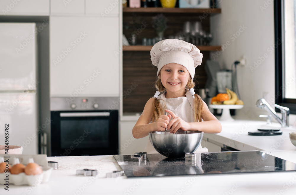 Small cute girl in special clothes and funny chef cap stay on kitchen and make dough for cake. Making cookie at home on kitchen, kid get fun enjoy cook. Child make dessert Small cute girl in special clothes and funny chef cap stay on kitchen and make dough for cake. Making cookie at home on kitchen, kid get fun enjoy cook. Child make dessert