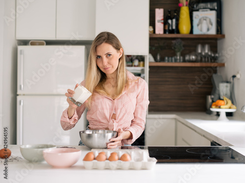 Cute blonde woman in pink blouse stays in kitchen, holds glass of flour, eggs are on table, going to make dough for cake. Person loves to cook. Girl prepares dessert according to delicious recipe.