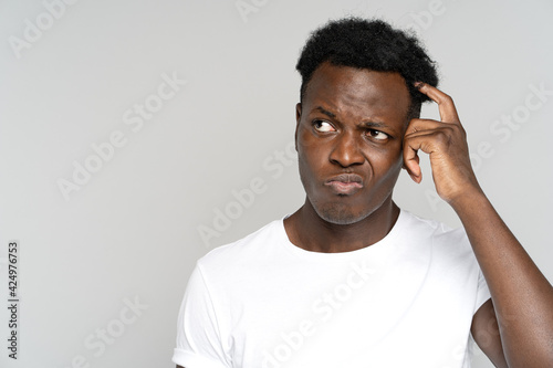 Studio portrait of unsure doubtful African man scratching head, looking left doubtfully at copy space, plans or reconsiders something, feeling hesitant poses against grey background. Suspicion concept photo
