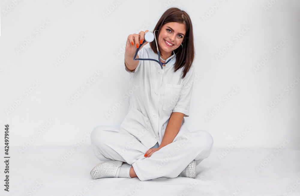 Young doctor in white uniform and clogs sitting on the floor with stethoscope. She is smiling. Isolated photography. Young doctor in white uniform and clogs sitting on the floor with stethoscope. She is smiling. Isolated photography.