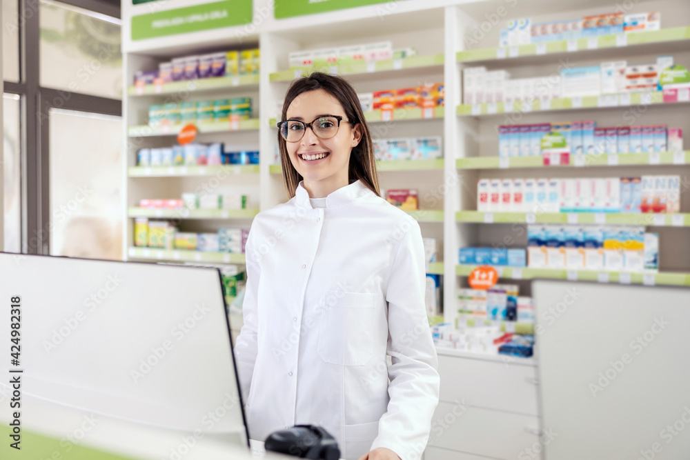 Pharmacist at the counter. A young woman in a white uniform focused on work at the computer is smiling at patients. The woman at work, pharmacist job, professional development