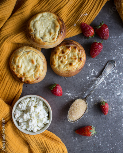 Authentic Russian open pies with cottage cheese, strawberry, ceramic cup with milk and bowl with cottage on a table. Yellow fabric background. Homemade pastries top view photo. 