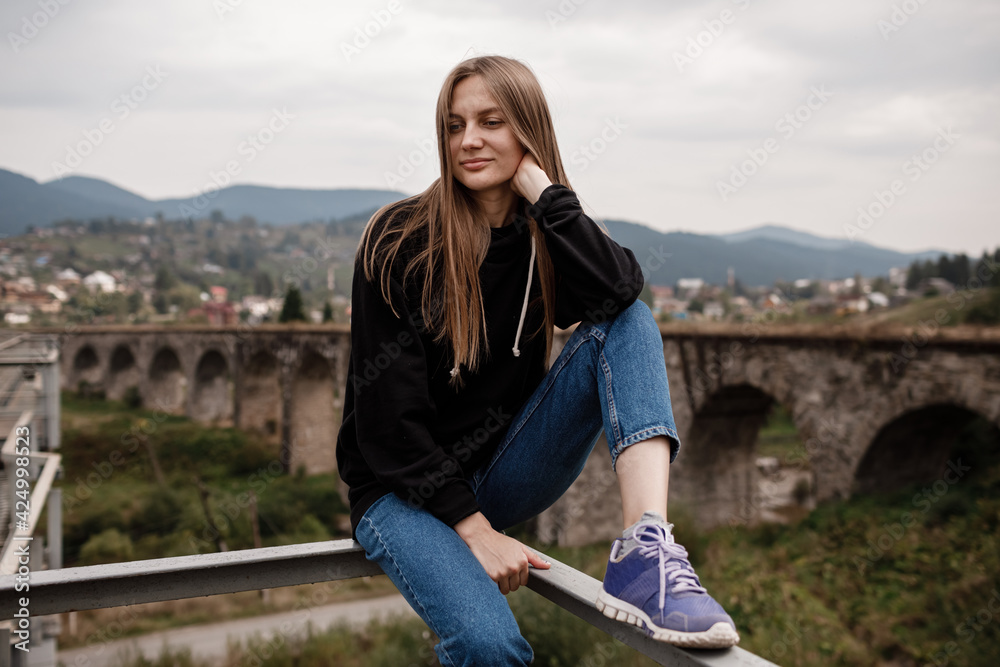 Naklejka premium Young woman is sitting near old viaduct. Tourist girl in scenery countryside by the Historical abandoned railway arch bridge viaduct in Vorokhta, Ivano-Frankivsk Region, Ukraine