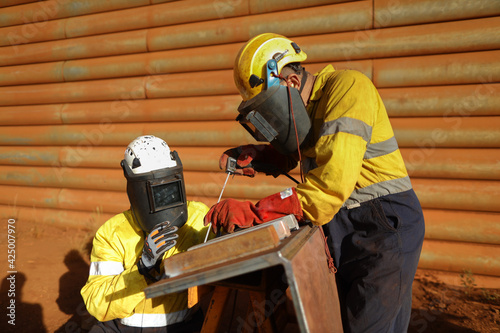 Safety workplace welder supervisor wearing dark shield mask welding PPE teaching guiding inspecting new worker on site while co worker welder is welding repairing chute liners Australia mining site