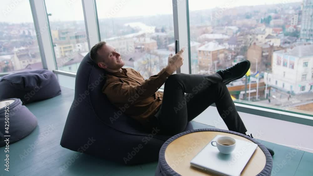Young businessman relaxing near the window. Handsome man sitting in a cosy chair and having a video conversation with the city window view. Profile view.