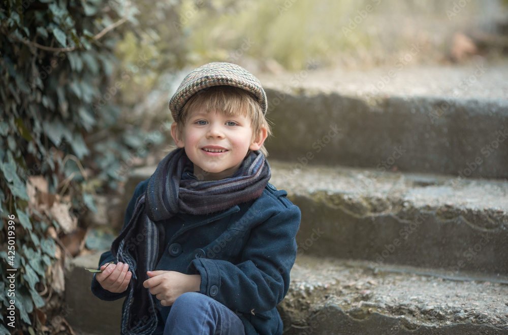 Portrait of a small child boy sitting on the steps and smiling with a leaf in his hand. Beautiful appearance of a bully, hipster or informal, vintage clothing.. Portrait of a small child boy sitting on the steps and smiling with a leaf in his hand. Beautiful appearance of a bully, hipster or informal, vintage clothing..