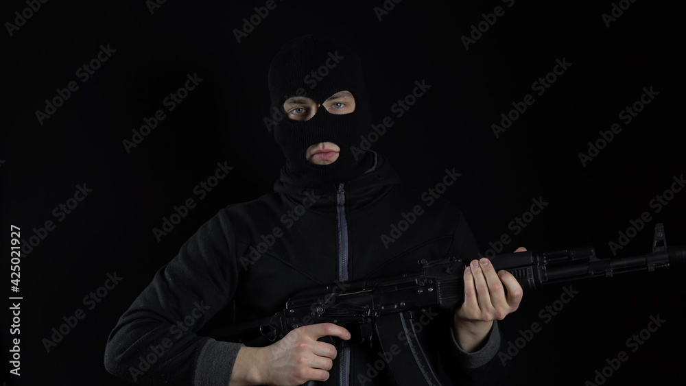 A man in a balaclava mask stands with an AK-47 assault rifle. The bandit charges the machine and stands. On a black background.