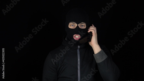 A man in a balaclava mask with a phone from the 90s. The bandit is talking on the phone. On a black background.