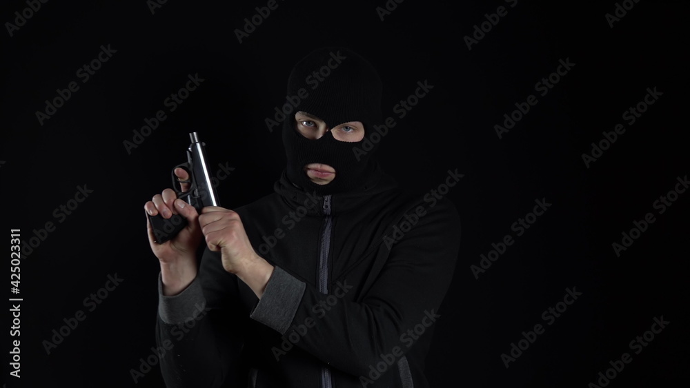 A man in a balaclava mask stands with a gun. The bandit reloaded the gun and stands on a black background. A man in a balaclava mask stands with a gun. The bandit reloaded the gun and stands on a black background.