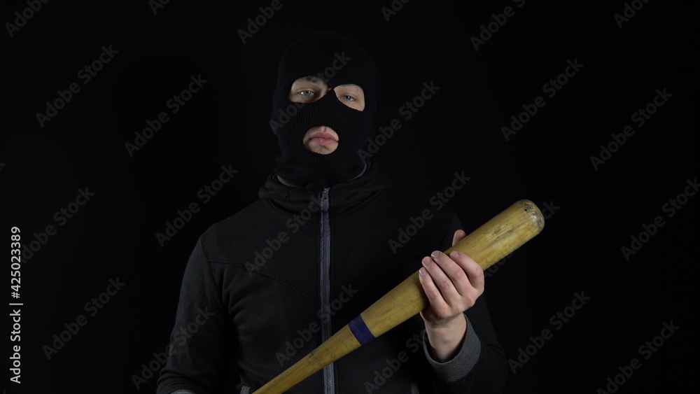 A man in a balaclava mask is standing with a baseball bat. A gangster is holding a baseball bat on a black background. A man in a balaclava mask is standing with a baseball bat. A gangster is holding a baseball bat on a black background.