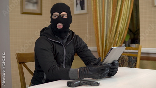 A robber is trying to hack into a tablet. A masked thug is sitting in a house and cannot hack into a tablet. Theft of data from a tablet.