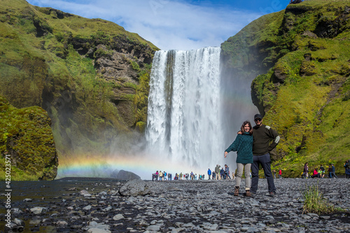 A couple at the Skogafoss waterfall on a summer afternoon with a rainbow and tourists under the waterfall. Iceland