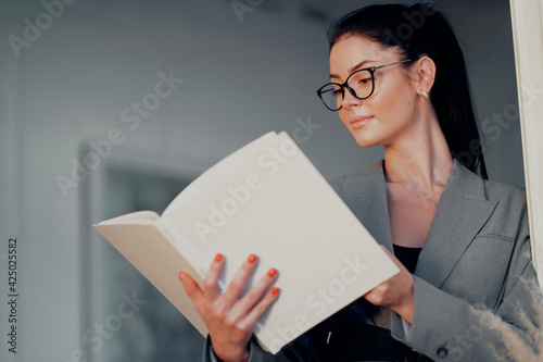 Smart female brunette student with glasses reading a book. Learns a foreign language from a textbook. Modern business suit.