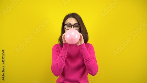 A young woman inflates a pink balloon with her mouth on a yellow background. Girl in a pink turtleneck and glasses.