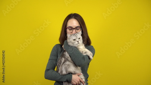 Young woman with a cat in her arms is dancing on a yellow background. Woman with glasses and a green sweater.