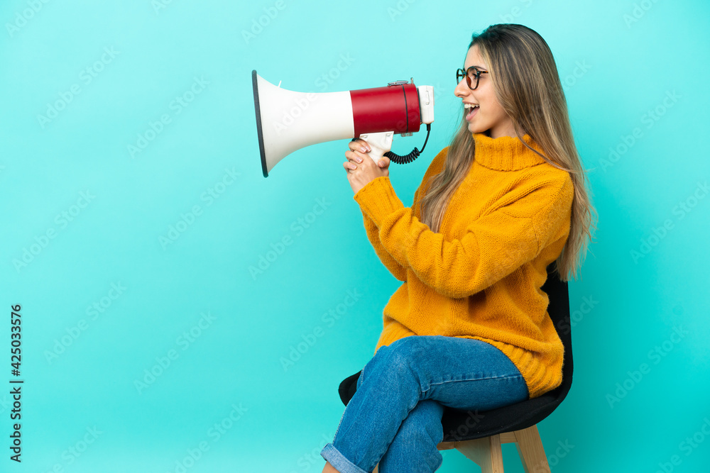 Young caucasian woman sitting on a chair isolated on blue background shouting through a megaphone Young caucasian woman sitting on a chair isolated on blue background shouting through a megaphone