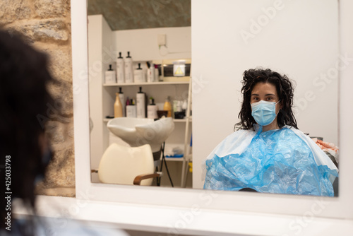 Young woman with mask looks to camera in hairdresser mirror