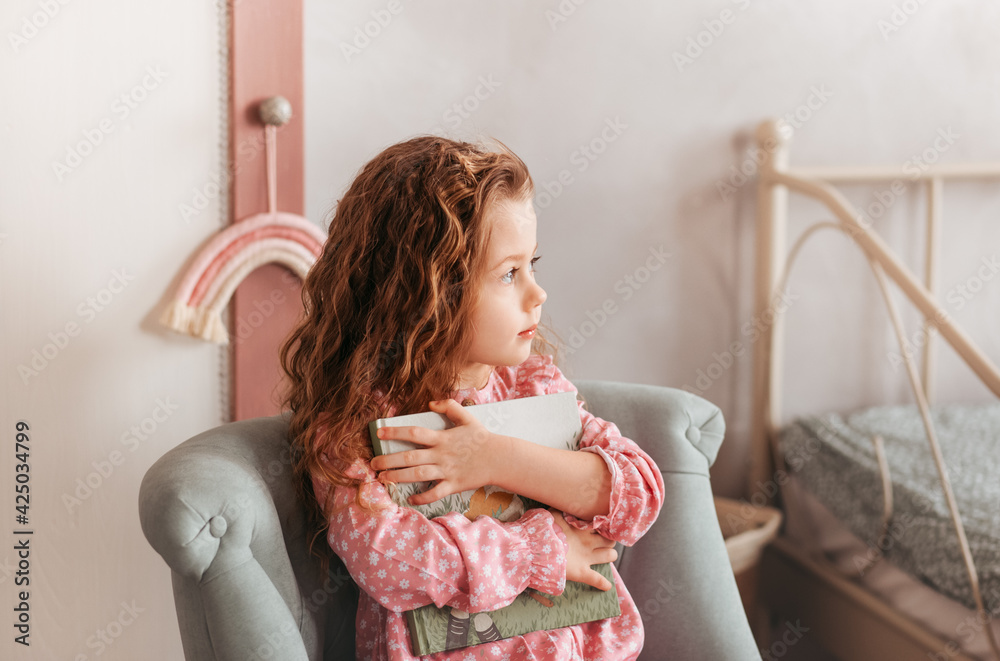 A little romantic girl with long hair sits in a chair and holds a book