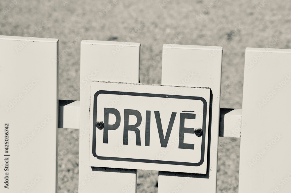 Wooden white fence closeup, a shield with the word privé (french for private) on it, creative black and white vintage style, blurred gravel path background, a sunny day Wooden white fence closeup, a shield with the word privé (french for private) on it, creative black and white vintage style, blurred gravel path background, a sunny day