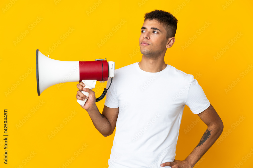 Young brazilian man isolated on yellow background holding a megaphone and thinking Young brazilian man isolated on yellow background holding a megaphone and thinking