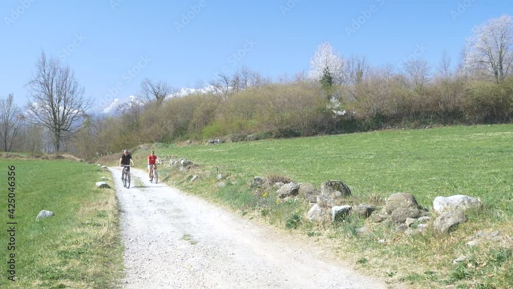Slow motion: Couple having fun by riding mountain bike on dirt road in sunny day, scenic landscape of snowcapped mountain range. Man and woman on bicycle in nature Slow motion: Couple having fun by riding mountain bike on dirt road in sunny day, scenic landscape of snowcapped mountain range. Man and woman on bicycle in nature
