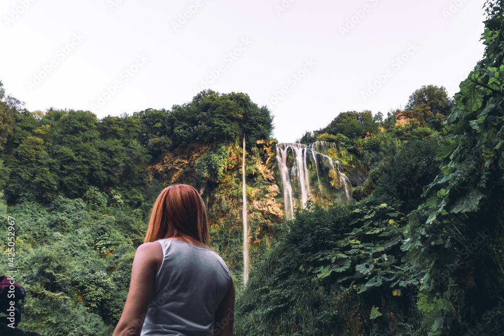 Naklejka premium Tourist girl pose, look at waterfall in Italy (Cascata delle Marmore) in Umbria. small, calm water pressure. Evening, sunset. Summer, bottom view