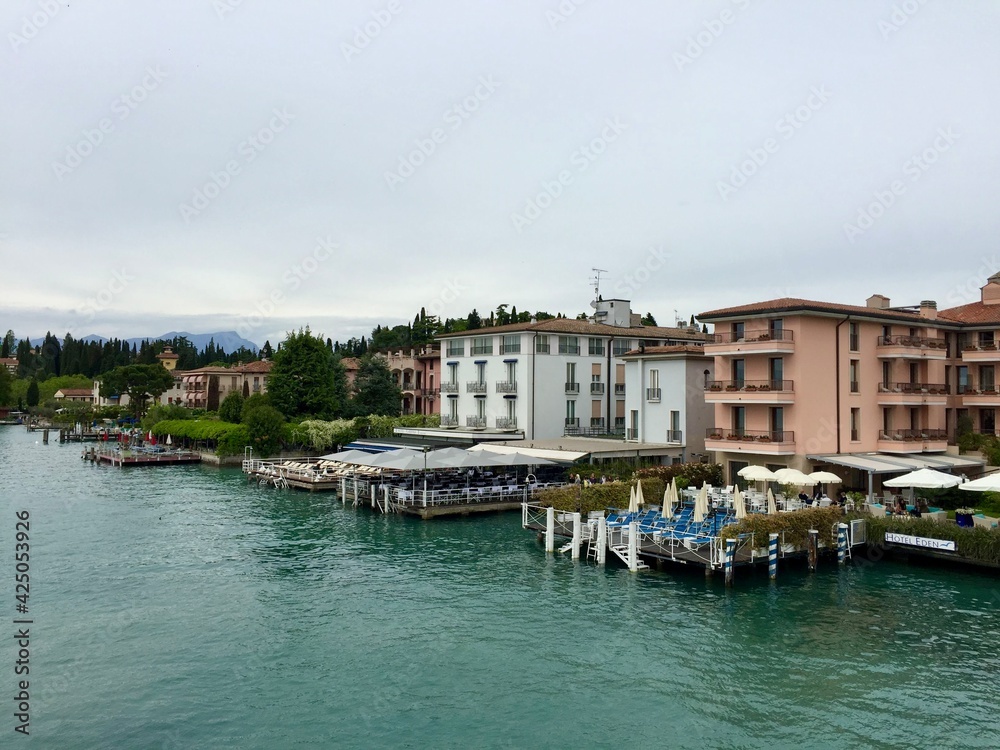 Approaching to Sirmione, Garda lake, Italy. Turquoise blue water. Beautiful colorful buildings around. Reflection in water. Cloudy weather.