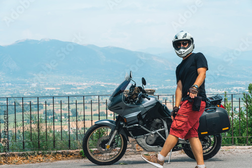 A lightly dressed man and his adventure motorcycle. No protection, only helmet. Sunny day in Narni, Italy. Green mountains in the background. Vacation and travel concept