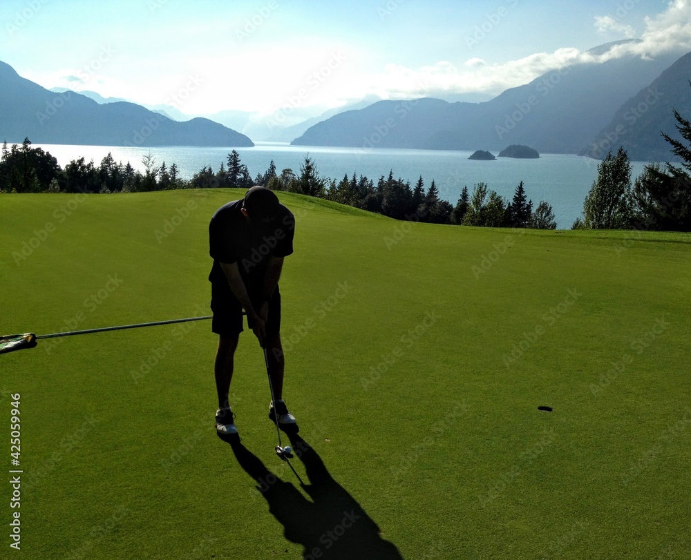 Obraz premium Man putting golf ball into hole with the ocean of Howe Sound British Columbia in the background. Very scenic