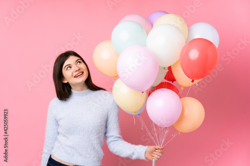 Young Ukrainian teenager girl holding lots of balloons over isolated pink background laughing and looking up