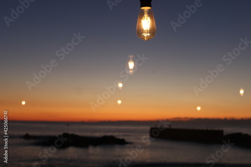 View from the window to the ocean at sunset. A photograph is a background where the main object is a burning light bulb and its multiple reflection in the window.