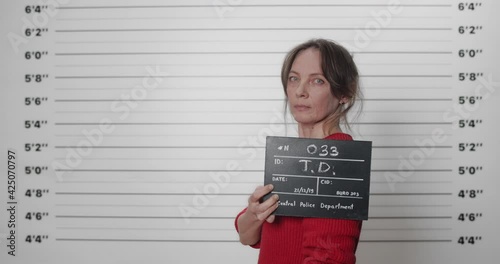 Side profle mugshot of woman in middle 40s turning head while holding sign and posing for photo. Portrait of female criminal looking to camera in police department at lineup metric wall. photo