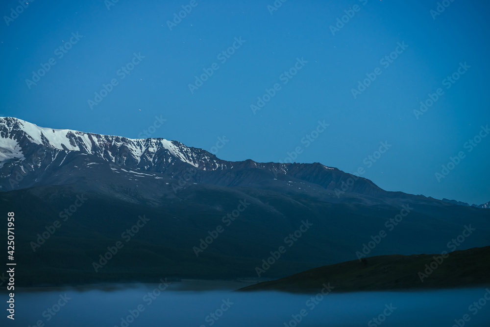 Atmospheric mountains landscape with dense fog and great snow mountain range under twilight sky. Alpine scenery with big snowy mountain ridge over thick fog in night. Snowy rocks above clouds in dusk.