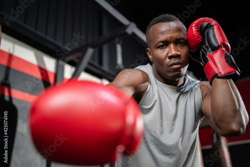 African American fighter man wear boxing red gloves and punching forward with one fist to camera in the fitness sport gym. Make looking forward with serious face. Exercise for a healthy body © bung