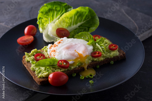 Fresh spring sandwich on a sliced gluten free sunflower bread. Red and green vegetables on black slate. Poached egg, avocado, chili and spring onion garnished with plum tomatoes