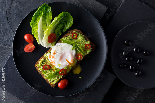 Fresh spring sandwich on a sliced gluten free sunflower bread. Red and green vegetables on black slate. Poached egg, avocado, chili and spring onion garnished with plum tomatoes  and black olives