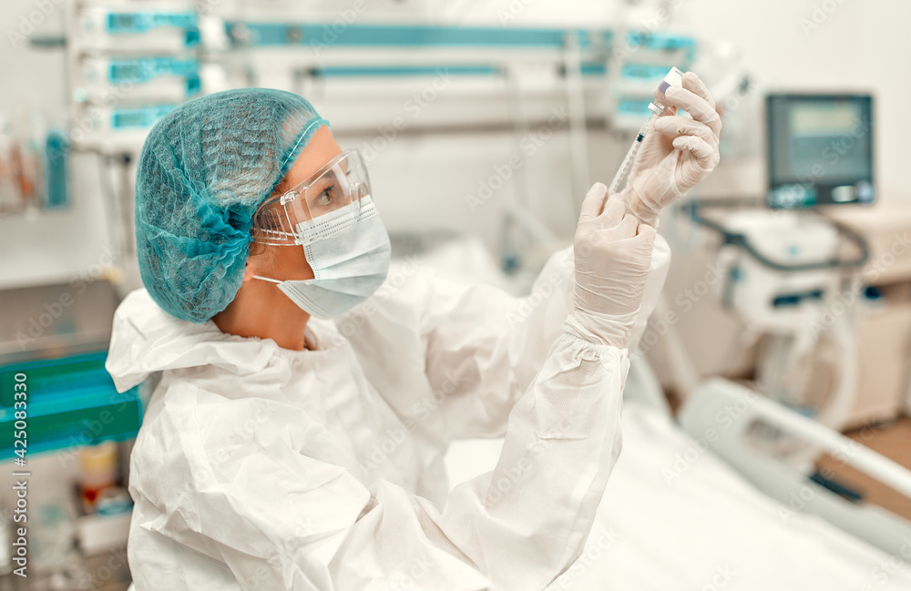 Fototapeta premium A doctor in a protective suit holds a syringe and an ampoule with the covid-19 coronavirus vaccine while standing in an intensive care unit in a modern hospital.